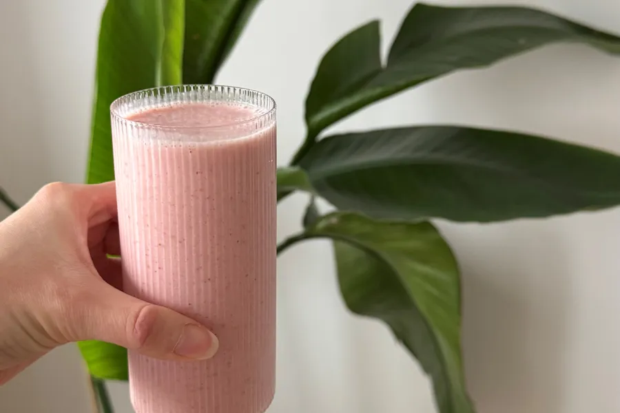 En hånd som holder et glass med rosa væske med en plante i bakgrunnen